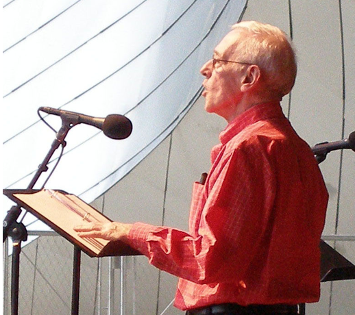 Photo of John Mahoney wearing a salmon colored shirt, talking into a microphone.