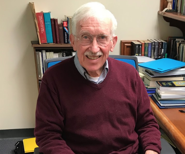 Photo features Dr. Falla, a man with gray hair wearing a maroon sweatshirt with a background of brightly colored books.