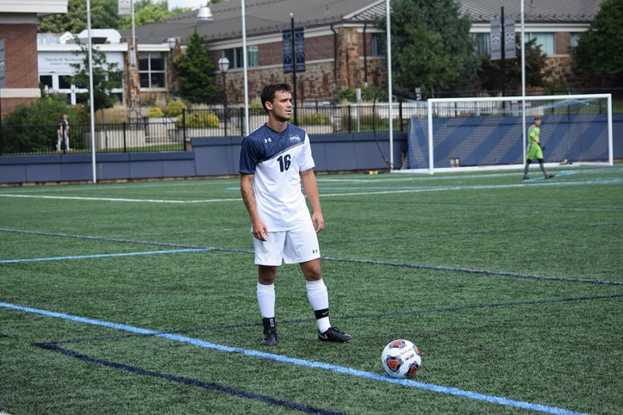 Mike Guarino is featured in a blue and white uniform on a lush green field waiting to kick a soccer ball.