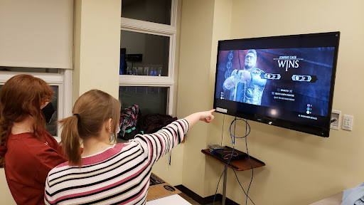 Abigail Schoepple, 23’, (right) teaches Gianna Tully, 24’, how to navigate the menus after finishing a fight in Mortal Kombat X during Game Night. Photo courtesy of Evan Yandrisovitz